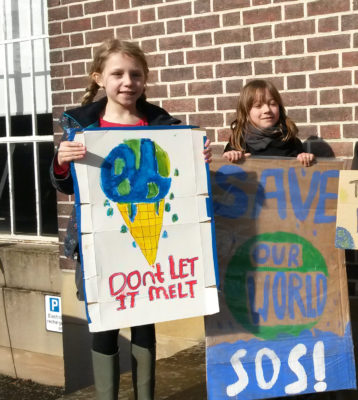 Two girls with placards reading 'don't let it melt' and 'save our world SOS' standing in front of a brick wall