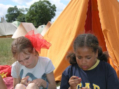 two children making friendship bands in front of a tent